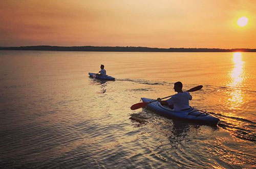 Two people kayaking on a calm lake during sunset, with the sun low in the sky and the water reflecting the warm orange light—a perfect evening adventure for guests staying at a Traverse City Bed and Breakfast.